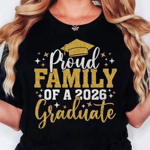 K&ouml;nnte beinhalten: Schwarzes T-Shirt mit dem Text "Proud Family of a 2026 Graduate" in wei&szlig;er und goldener Glitzer-Schrift. Eine goldene Abschlusskappe-Grafik befindet sich &uuml;ber dem Text. Das Shirt hat einen Rundhalsausschnitt.