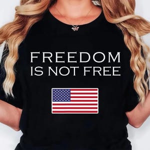 May include: A black short-sleeved t-shirt featuring the white text FREEDOM IS NOT FREE printed in a bold, sans-serif font. Below the text is a rectangular graphic of the United States flag with its traditional red, white, and blue colors.