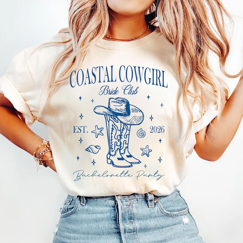 May include: A cream-coloured t-shirt featuring a blue graphic. The graphic includes the text "COASTAL COWGIRL Bride Club" above a drawing of a cowboy hat and boots. Below the graphic are the words "EST. 2026" and "Bachelorette Party".