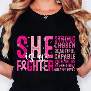 May include: Black t-shirt with pink text that reads "SHE is a FIGHTER" and other positive affirmations. The design includes a pink ribbon, flowers, and stars. The shirt is a symbol of strength and support.