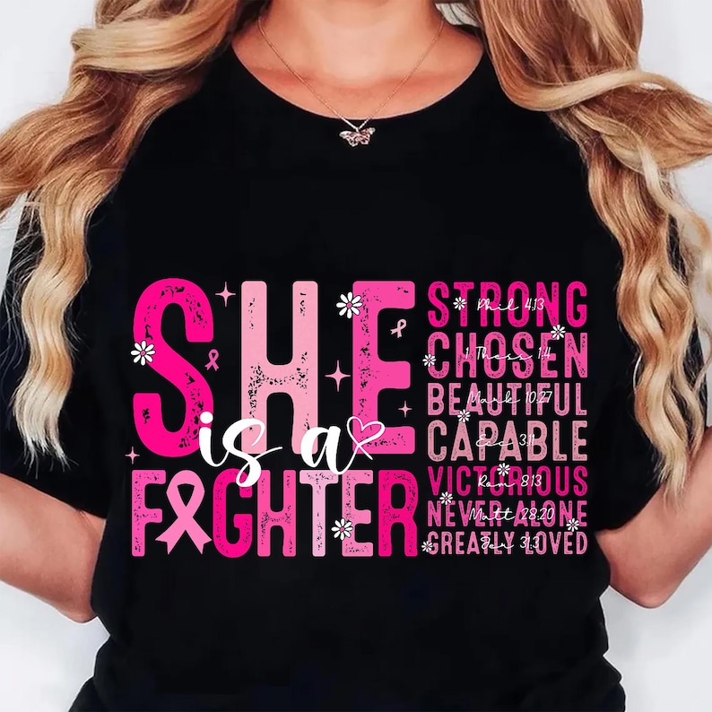 May include: Black t-shirt with pink text that reads "SHE is a FIGHTER" and other positive affirmations. The design includes a pink ribbon, flowers, and stars. The shirt is a symbol of strength and support.