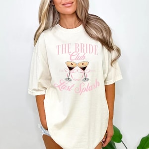 May include: A cream-coloured t-shirt featuring the pink text "THE BRIDE Club" and a graphic of two martini glasses. Below the graphic is the text "Last Splash". The shirt has short sleeves and a relaxed fit.