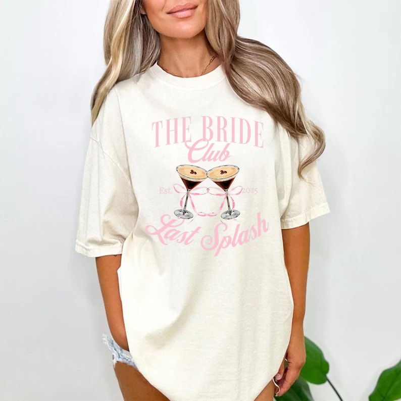 May include: A cream-coloured t-shirt featuring the pink text "THE BRIDE Club" and a graphic of two martini glasses. Below the graphic is the text "Last Splash". The shirt has short sleeves and a relaxed fit.