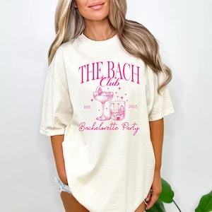 May include: A cream-coloured t-shirt featuring a pink graphic. The text "THE BACH Club" is above a cocktail glass and a rocks glass. Below the glasses, the words "Bachelorette Party" are displayed. The year "2025" is also printed on the shirt.
