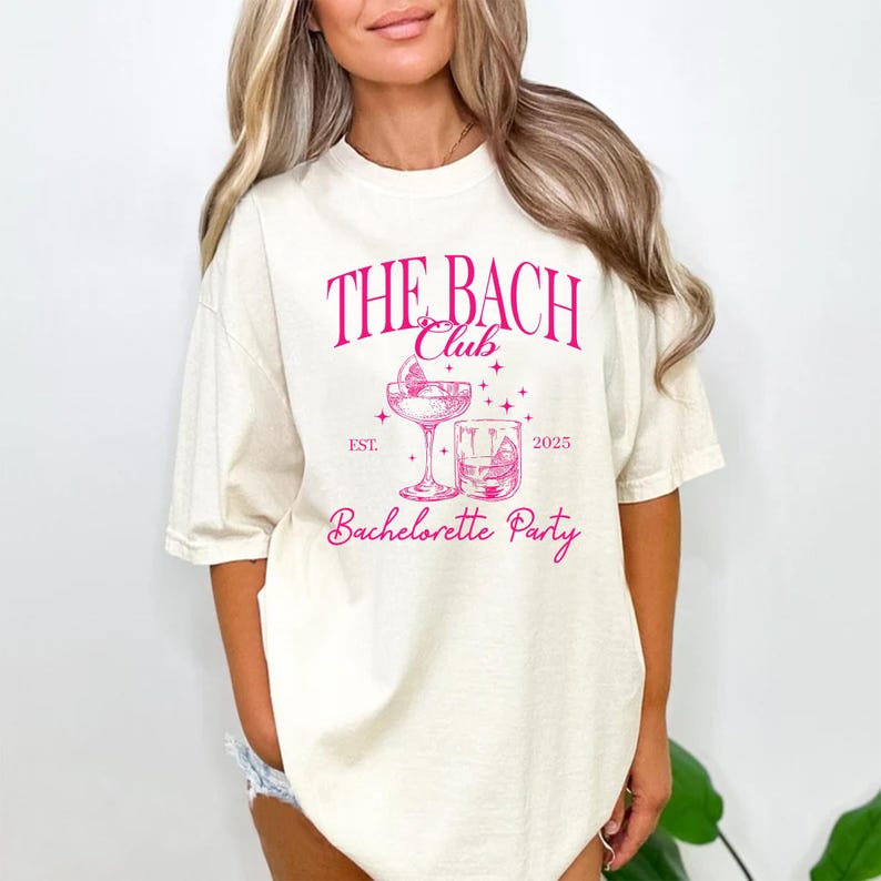 May include: A cream-coloured t-shirt featuring a pink graphic. The text "THE BACH Club" is above a cocktail glass and a rocks glass. Below the glasses, the words "Bachelorette Party" are displayed. The year "2025" is also printed on the shirt.