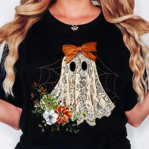 May include: Black t-shirt featuring a whimsical ghost design. The ghost is adorned with a brown bow and a patterned sheet, with floral accents in white, green, and orange. A spiderweb design is in the background.