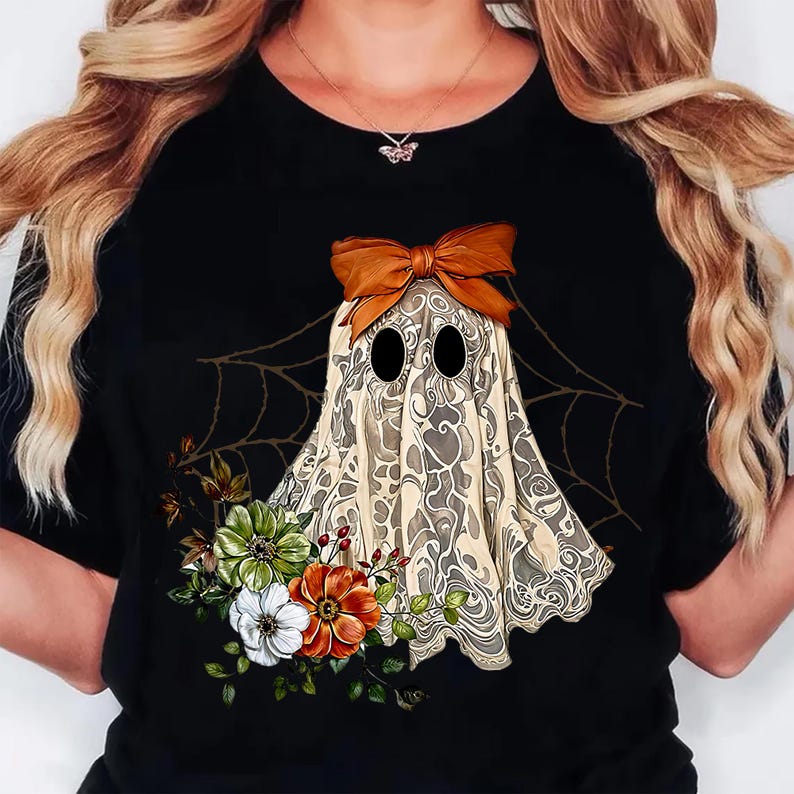 May include: Black t-shirt featuring a whimsical ghost design. The ghost is adorned with a brown bow and a patterned sheet, with floral accents in white, green, and orange. A spiderweb design is in the background.