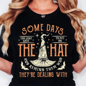 May include: Black t-shirt with orange text and a witch hat graphic. The text reads "SOME DAYS YOU JUST HAVE TO PUT ON THE HAT AND REMIND THEM WHO THEY'RE DEALING WITH." The witch hat has a spider web design.