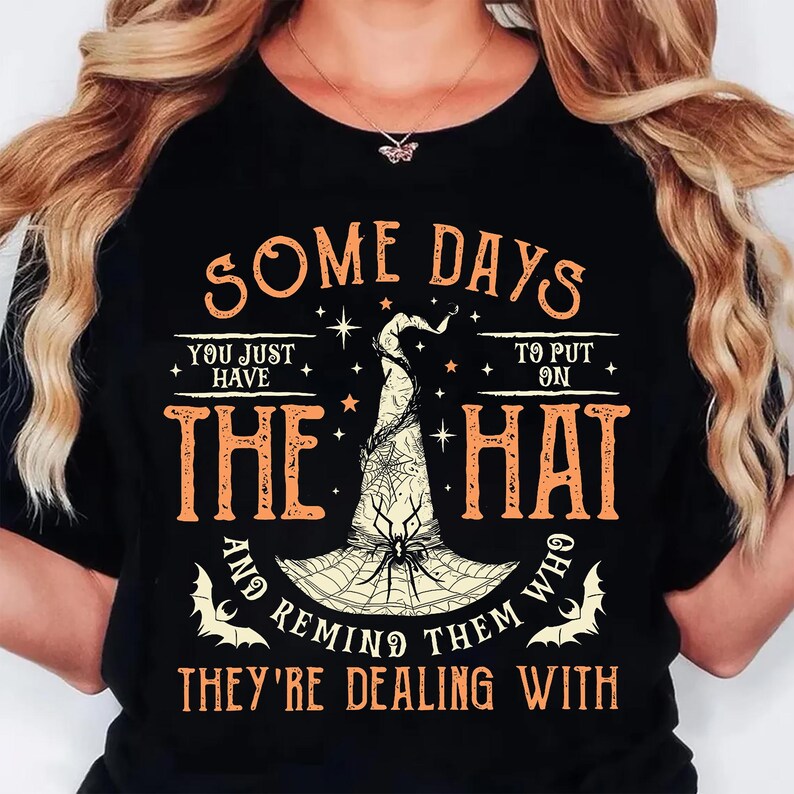 May include: Black t-shirt with orange text and a witch hat graphic. The text reads "SOME DAYS YOU JUST HAVE TO PUT ON THE HAT AND REMIND THEM WHO THEY'RE DEALING WITH." The witch hat has a spider web design.