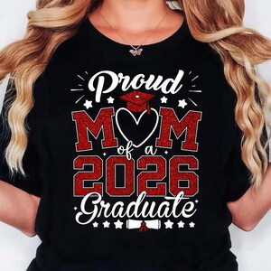 K&ouml;nnte beinhalten: Schwarzes T-Shirt mit einem roten Glitzer-Design mit der Aufschrift "Proud Mom of a 2026 Graduate". Das Design umfasst eine Abschlusskappe, Sterne und das Wort "Mom" in einem Herzen.