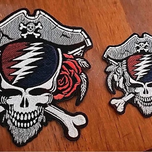 May include: Two embroidered patches featuring a skull wearing a pirate hat, with crossbones and roses. The skull has a red, white, and blue design. One patch is larger than the other.