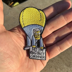 May include: Embroidered patch featuring a cartoon character in a yellow hot air balloon. The character is wearing a blue suit and holding a sign that reads "Hi I'm Big Butt Skinner". The patch has a black border.