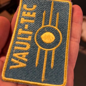 Puede incluir: Un parche rectangular con el texto "VAULT-TEC" en letras doradas. El parche tiene un fondo verde azulado con un emblema dorado en el centro. El emblema es un círculo con un ojo estilizado y tres líneas verticales.