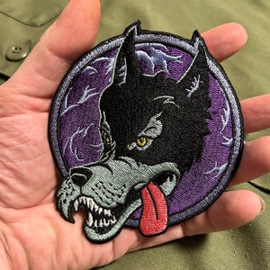 Op de afbeelding: Ronde geborduurde patch met een wolfskop met een rode tong, tegen een paarse achtergrond met een gestileerd ontwerp. De wolf heeft zwarte vacht, een grijze snuit en gele ogen.
