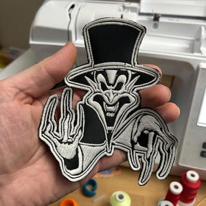 May include: A black and white embroidered patch depicting a figure in a top hat and cape. The patch has detailed stitching and a spooky, cartoonish design. The figure's hands are outstretched, and the overall aesthetic is reminiscent of vintage horror or Halloween themes.