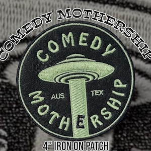 Puede incluir: Parche bordado en negro y verde con las palabras "COMEDY MOTHERSHIP" en una fuente curva. El parche presenta un diseño de OVNI verde con las palabras "COMEDY" y "MOTHERSHIP" en verde. También es visible el texto "AUS TEX". El parche mide 10 cm.