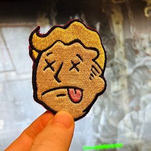 May include: Embroidered patch featuring a cartoon face with crossed-out eyes and a lolling tongue. The patch has a tan face, blonde hair, and a black outline. The patch is being held up by a hand.