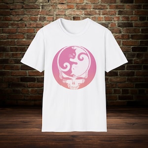 May include: White t-shirt featuring a pink and white graphic design. The design is a stylized skull with a yin-yang symbol in the center, set within a pink circle. The design has a striped pattern.