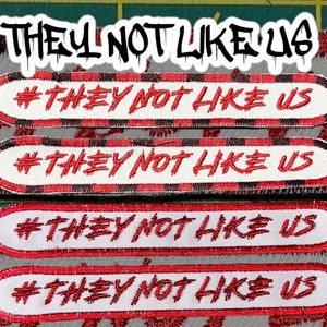 Op de afbeelding: Vier geborduurde patches met de tekst "#THEY NOT LIKE US" in rood op een witte achtergrond. Elke patch heeft een rode rand, en de bovenste twee hebben een zwart-rode geblokte rand. De zin "THEY NOT LIKE US" staat boven de patches.