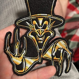 May include: Embroidered patch depicting a grinning figure in a top hat, using black, gold, and white thread. The patch has a detailed outline, suitable for application on clothing or accessories. The design is likely for decorative purposes.