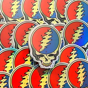 May include: A collection of colorful stickers featuring a stylized skull with a lightning bolt design. The stickers have a red and blue background with a yellow lightning bolt and a silver border. The stickers are holographic.