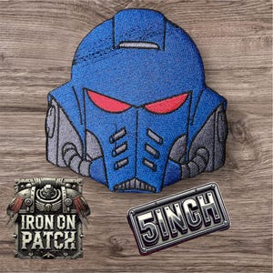 May include: A blue embroidered patch shaped like a futuristic helmet with red eyes. A rectangular metal badge reads "5 INCH" (approximately 12.7 cm), and a square patch says "IRON ON PATCH".