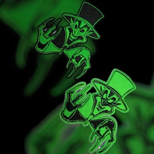 May include: Two embroidered patches featuring a green and black cartoon character with a top hat and clawed hands. The patches have a glow-in-the-dark effect against a black background. The design is repeated twice.
