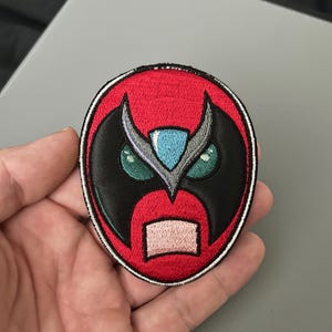 以下が含まれることがあります： スタイリッシュなマスクデザインの刺繍パッチ。パッチは赤い背景に黒のアクセント、白い縁取り、緑の目、青い宝石、ピンクの口を持つ詳細な顔が特徴です。パッチは丸いです。