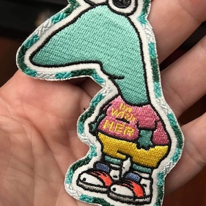 May include: Embroidered patch featuring a cartoon dinosaur with a teal head and a pink shirt that says "I'm with HER". The dinosaur wears yellow shorts and red sneakers. The patch has a white border with green accents.