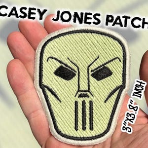 May include: Embroidered patch featuring the mask of Casey Jones, a character from the Teenage Mutant Ninja Turtles. The patch is light green with black details and a white border. The patch measures 7.6cm x 9.7cm and has the text "CASEY JONES PATCH".