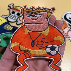 May include: An embroidered patch featuring a cartoon character with spiky yellow hair, a peach-coloured face, and a sceptical expression. The character wears an orange shirt with a yellow collar and a black whistle, holding a black and white football.