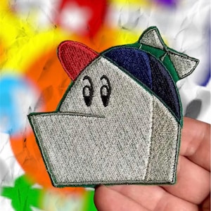 May include: Embroidered patch featuring a cartoon character with a silver helmet, red visor, blue cap, and green bow. The character has black eyes and a smiling expression. The patch is held against a colorful, abstract background.