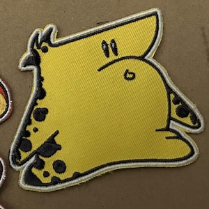 May include: A yellow embroidered patch featuring a cartoon giraffe design. The giraffe has black spots and a black outline. The patch is on a brown surface.