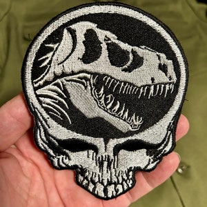 以下が含まれることがあります： 黒と白の刺繍パッチ。恐竜の頭蓋骨のデザインが、頭蓋骨の形をしたアウトラインの中にあります。このパッチは、衣類やアクセサリー用で、詳細で質感のある外観をしています。