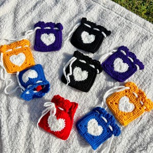 May include: Eight crocheted drawstring bags with white heart designs. The bags are in various colors, including purple, black, yellow, red, and blue.