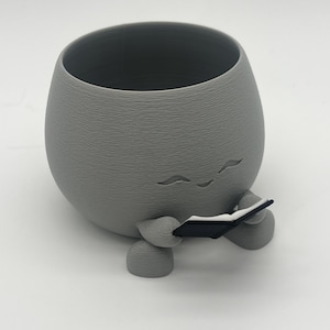 Op de afbeelding: Een grijze, eivormige plantenbak met een textuuroppervlak. De plantenbak heeft kleine voetjes en houdt een klein zwart boek vast. De plantenbak heeft een grillig ontwerp, met drie gebogen lijnen aan de bovenkant.