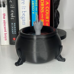 Puede incluir: Un caldero negro con tres patas, que contiene una mano gris con el dedo coraz&oacute;n levantado. El caldero est&aacute; frente a una estanter&iacute;a con libros, incluyendo "A Court of Thorns and Roses".