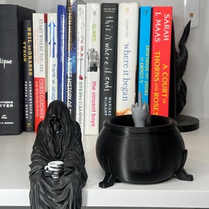 Puede incluir: Una figura negra de la Parca sosteniendo una taza de caf&eacute; se encuentra junto a un caldero negro con una mano haciendo una peineta. Hay libros en el fondo, incluyendo t&iacute;tulos como "A Court of Thorns and Roses" y "Where it began".