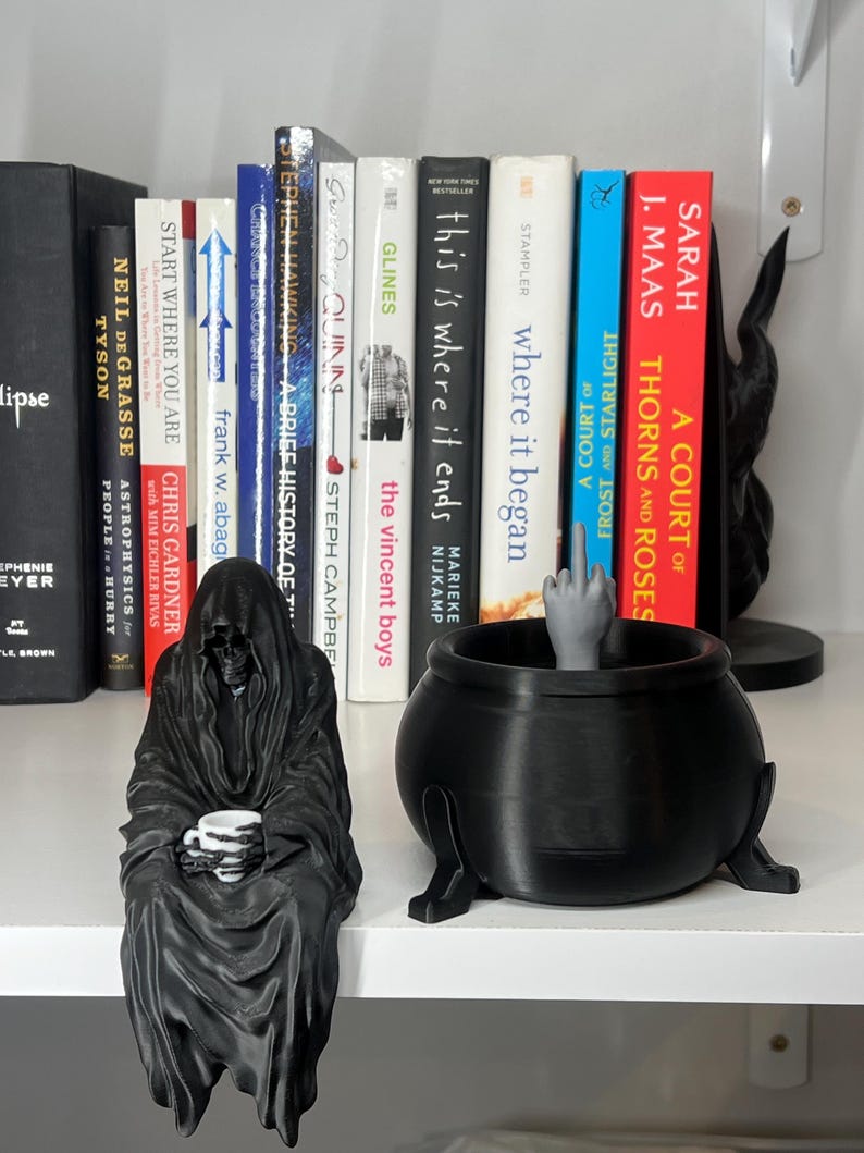 Puede incluir: Una figura negra de la Parca sosteniendo una taza de caf&eacute; se encuentra junto a un caldero negro con una mano haciendo una peineta. Hay libros en el fondo, incluyendo t&iacute;tulos como "A Court of Thorns and Roses" y "Where it began".