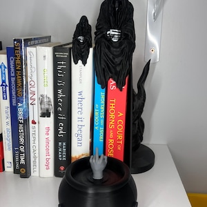 Puede incluir: Un caldero negro con un dedo medio gris y sujetalibros de la Parca en una estanter&iacute;a con libros. Los libros incluyen t&iacute;tulos como "A Court of Thorns and Roses" y "This is Where it Ends."