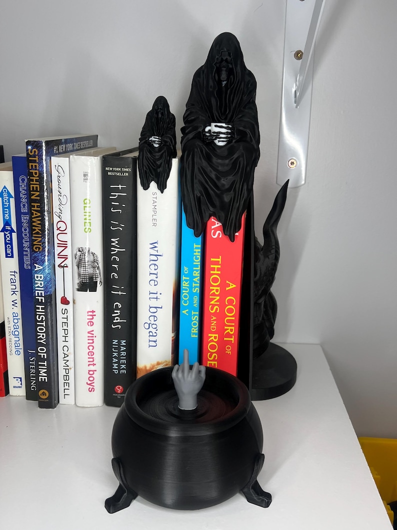 Puede incluir: Un caldero negro con un dedo medio gris y sujetalibros de la Parca en una estanter&iacute;a con libros. Los libros incluyen t&iacute;tulos como "A Court of Thorns and Roses" y "This is Where it Ends."