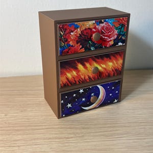 Puede incluir: Un peque&ntilde;o cofre de madera marr&oacute;n con tres cajones. Cada frente de caj&oacute;n presenta un dise&ntilde;o diferente: un estampado floral, llamas y un cielo nocturno con una luna creciente y estrellas. Los pomos son redondos y de madera.