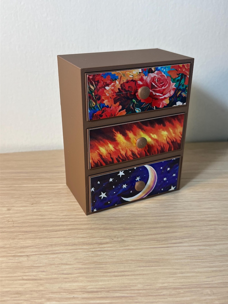 Puede incluir: Un peque&ntilde;o cofre de madera marr&oacute;n con tres cajones. Cada frente de caj&oacute;n presenta un dise&ntilde;o diferente: un estampado floral, llamas y un cielo nocturno con una luna creciente y estrellas. Los pomos son redondos y de madera.