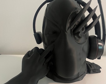May include: A black 3D printed figurine of a masked figure wearing headphones. The figure has a hand raised in a gesture of defiance. The figurine is designed to hold headphones.