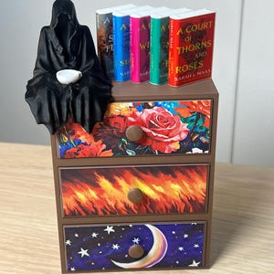 Puede incluir: Una peque&ntilde;a c&oacute;moda marr&oacute;n con tres cajones, cada uno decorado con diferentes dise&ntilde;os. Una figura negra con capucha est&aacute; sentada encima, sosteniendo una taza blanca. Libros con lomos coloridos est&aacute;n dispuestos en la parte superior. Los cajones presentan patrones florales, de fuego y celestiales.