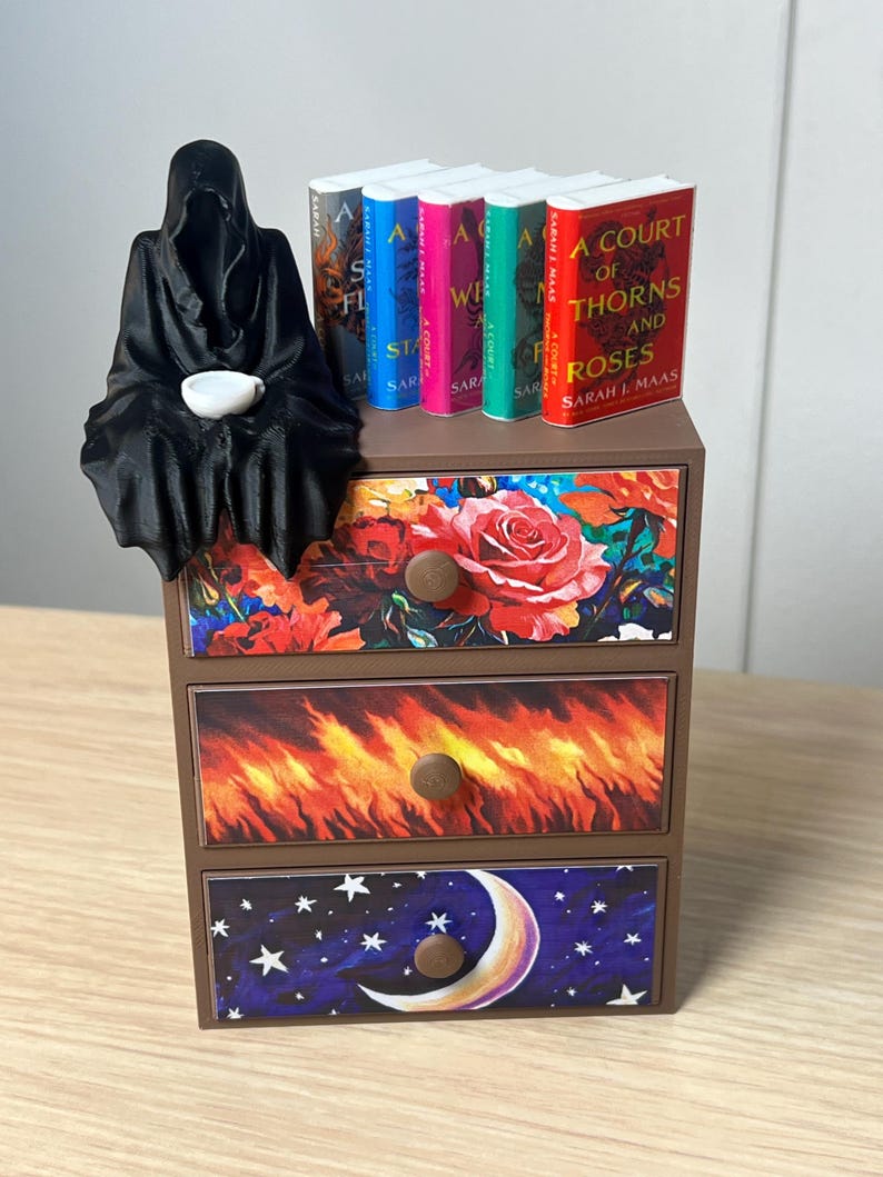 Puede incluir: Una peque&ntilde;a c&oacute;moda marr&oacute;n con tres cajones, cada uno decorado con diferentes dise&ntilde;os. Una figura negra con capucha est&aacute; sentada encima, sosteniendo una taza blanca. Libros con lomos coloridos est&aacute;n dispuestos en la parte superior. Los cajones presentan patrones florales, de fuego y celestiales.