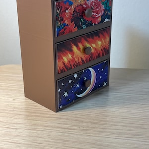 Puede incluir: Un peque&ntilde;o cofre marr&oacute;n de tres cajones con frentes decorativos. El caj&oacute;n superior presenta un dise&ntilde;o floral con rosas rojas, el caj&oacute;n central tiene un patr&oacute;n de fuego y el caj&oacute;n inferior muestra un cielo nocturno con una luna creciente y estrellas. Cada caj&oacute;n tiene un pomo redondo marr&oacute;n.