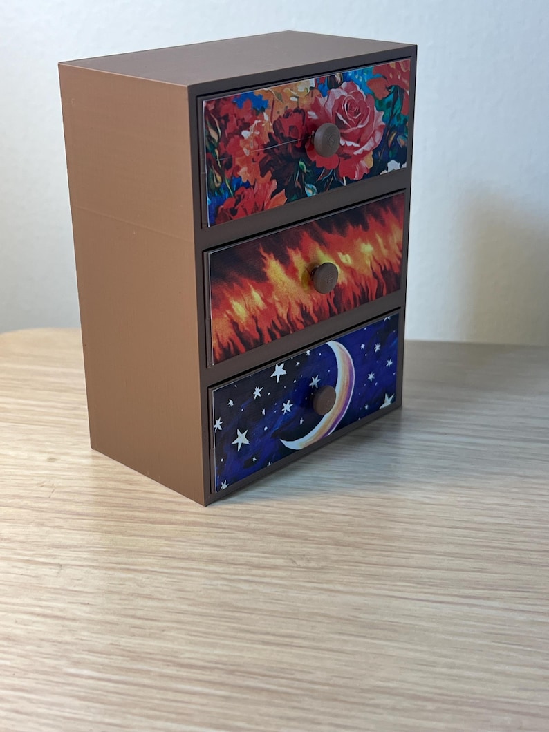 Puede incluir: Un peque&ntilde;o cofre marr&oacute;n de tres cajones con frentes decorativos. El caj&oacute;n superior presenta un dise&ntilde;o floral con rosas rojas, el caj&oacute;n central tiene un patr&oacute;n de fuego y el caj&oacute;n inferior muestra un cielo nocturno con una luna creciente y estrellas. Cada caj&oacute;n tiene un pomo redondo marr&oacute;n.