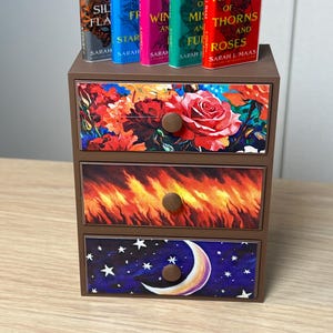 Puede incluir: Una peque&ntilde;a c&oacute;moda de madera marr&oacute;n con tres cajones, cada uno decorado con un dise&ntilde;o diferente. El caj&oacute;n superior presenta un estampado floral, el caj&oacute;n central un dise&ntilde;o de fuego y el caj&oacute;n inferior una escena de cielo nocturno. Cinco libros en miniatura encima.