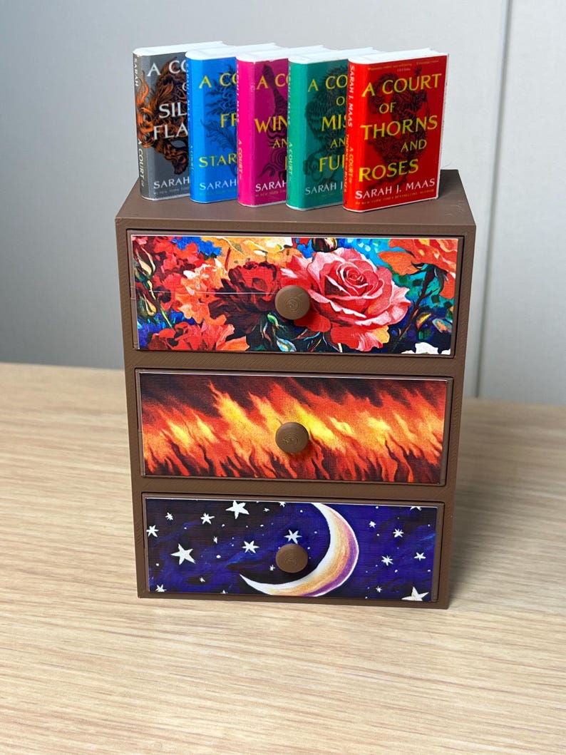 Puede incluir: Una peque&ntilde;a c&oacute;moda de madera marr&oacute;n con tres cajones, cada uno decorado con un dise&ntilde;o diferente. El caj&oacute;n superior presenta un estampado floral, el caj&oacute;n central un dise&ntilde;o de fuego y el caj&oacute;n inferior una escena de cielo nocturno. Cinco libros en miniatura encima.